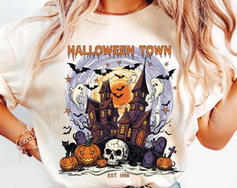 May include: Cream-colored t-shirt featuring a Halloween-themed graphic. The design includes a haunted house, bats, ghosts, pumpkins, and a skull. The text "HALLOWEEN TOWN" is arched across the top, with "EST. 1998" at the bottom.