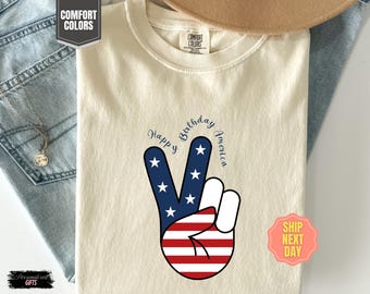 Peut inclure: T-shirt crème avec un motif de signe de paix aux couleurs du drapeau américain. Le signe de paix est composé d'étoiles bleues et blanches et de rayures rouges et blanches. Les mots "Happy Birthday America" sont arqués au-dessus.