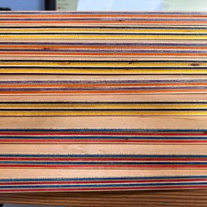 May include: Close-up of a skateboard deck made of multiple layers of wood with alternating colored plies. The plies are visible on the edge of the deck, showing a variety of colors including red, blue, yellow, and black.