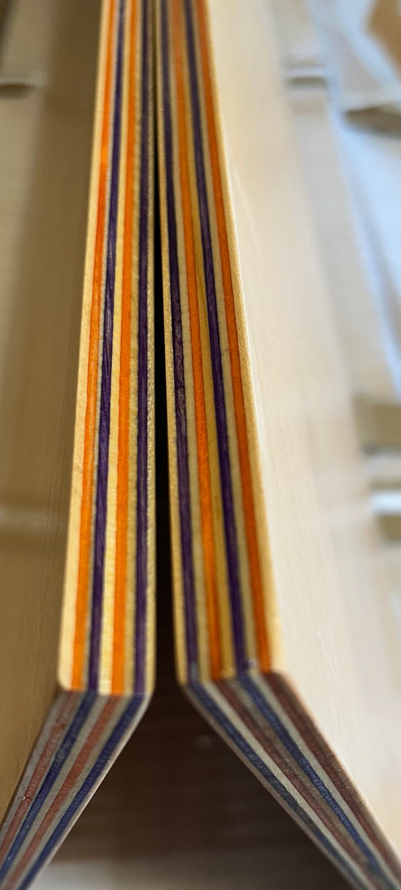 May include: Close-up of the edge of a wooden board, showing the layers of wood veneer in orange, purple, and brown.