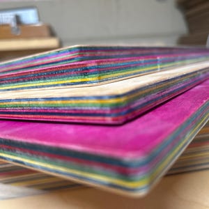 May include: Close-up of several colorful skateboard decks stacked on top of each other. The decks have a layered construction with visible stripes of various colors, including pink, blue, yellow, and green. The edges are smooth and rounded.