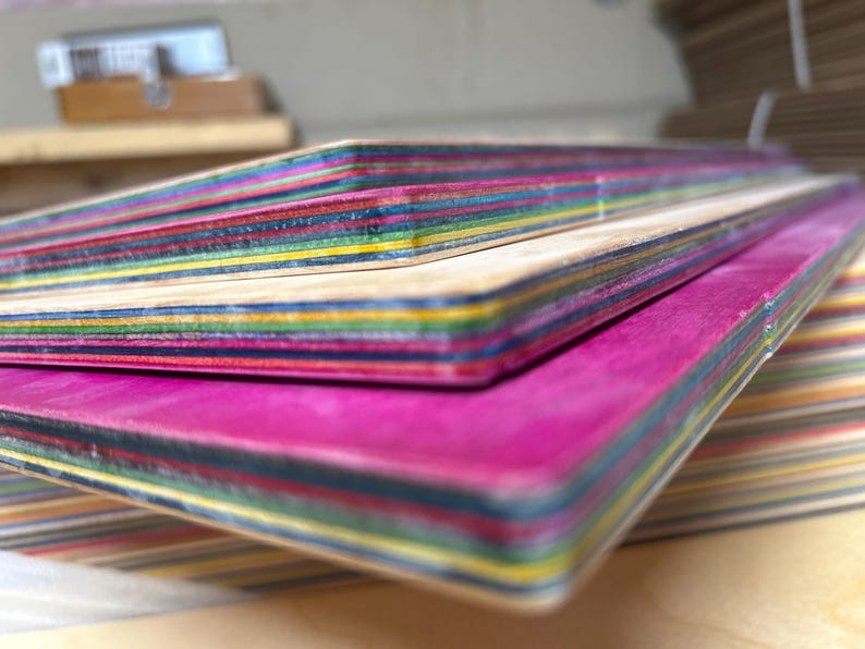 May include: Close-up of several colorful skateboard decks stacked on top of each other. The decks have a layered construction with visible stripes of various colors, including pink, blue, yellow, and green. The edges are smooth and rounded.