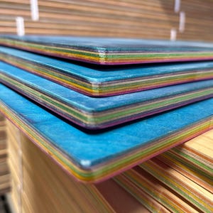 May include: Close-up of several blue skateboards stacked on top of each other. The edges of the boards display a rainbow of colors, and the top surface has a textured blue finish. The boards are made of layered wood.