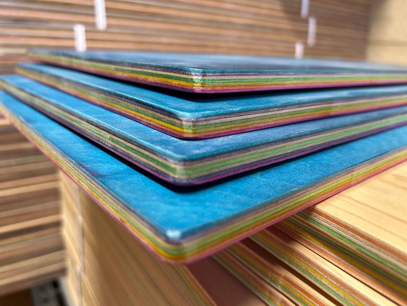 May include: Close-up of several blue skateboards stacked on top of each other. The edges of the boards display a rainbow of colors, and the top surface has a textured blue finish. The boards are made of layered wood.
