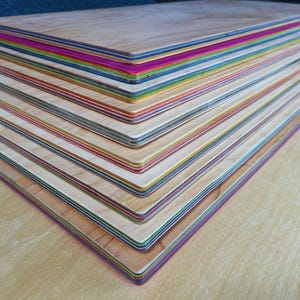 May include: A pile of wooden boards with edges in a rainbow of colours. The boards are made of light-coloured wood, with edges painted in various colours, including pink, blue, green, and yellow. The boards are stacked on top of each other.