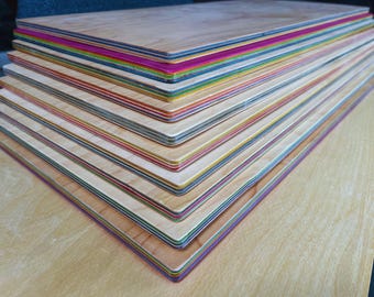 May include: A stack of wooden boards with colorful edges. The boards are made of light-colored wood and have edges painted in various colors, including pink, blue, green, and yellow. The boards are stacked on top of each other.