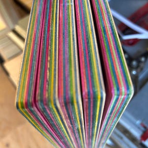 May include: Close-up of a stack of colorful, layered wood pieces. The edges reveal a vibrant array of colors including pink, yellow, green, and blue. The wood appears to be for a craft project or woodworking. The pieces are held together.