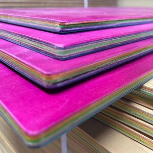 May include: Close-up of a stack of colorful wooden boards. The top surfaces are a vibrant magenta, with edges showing layers of various colors. The boards appear to be new and unused, with a smooth finish.