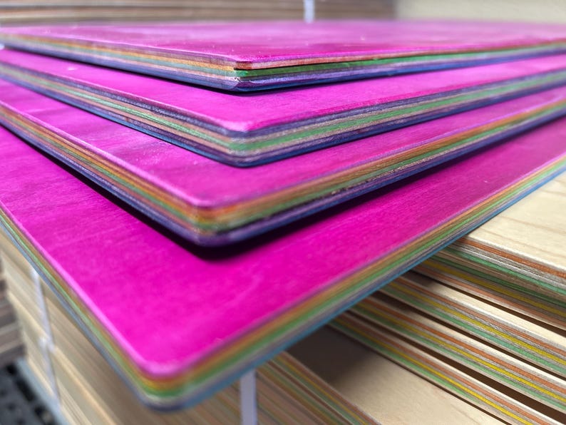 May include: Close-up of a stack of colorful wooden boards. The top surfaces are a vibrant magenta, with edges showing layers of various colors. The boards appear to be new and unused, with a smooth finish.