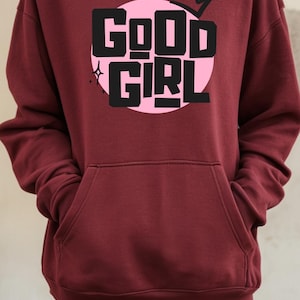 Może przedstawiać: Bordowa bluza z kapturem z różowym kołem graficznym, na którym widnieje napis "GOOD GIRL" czarnym tekstem z koroną nad tekstem.