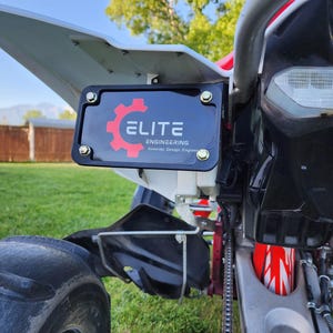 Puede incluir: Primer plano de un quad negro y rojo con el texto "ELITE ENGINEERING Innovate. Design. Engineer." El quad tiene un soporte de matrícula negro, un guardabarros blanco y un amortiguador rojo y blanco. El fondo es un césped verde y un cielo azul.