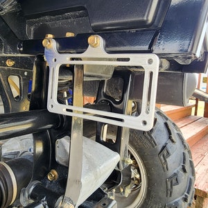 Puede incluir: Un marco plateado para la placa de matrícula está montado en un ATV negro. El marco es rectangular con esquinas redondeadas y se fija con pernos dorados. El ATV tiene una carrocería negra y un neumático negro grande.