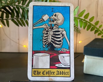 May include: A tarot card with a blue background and a skeleton drinking coffee. The card is titled "The Coffee Addict".