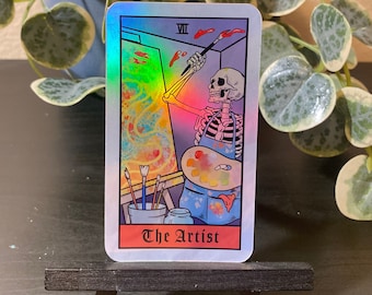 May include: A holographic tarot card with a skeleton painting on a canvas. The card is titled "The Artist" and has the Roman numeral VII in the top right corner.
