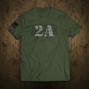 Pode incluir: Uma camiseta verde com uma bandeira americana branca na manga e uma citaat da Segunda Emenda da Constituição dos Estados Unidos impressa na frente. A citaat diz: "Uma milícia bem regulamentada, sendo necessária para a segurança de um Estado livre, o direito do povo de possuir e portar armas, não será violado."