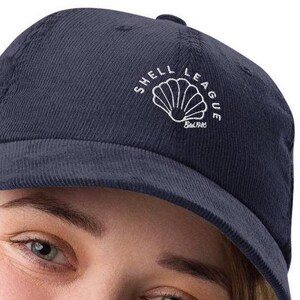 May include: A navy blue corduroy baseball cap with a white embroidered logo that says "Shell League" and features a seashell design. The logo also includes the text "Salt Life".