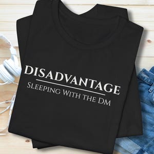 Puede incluir: Camiseta negra con el texto "DISADVANTAGE SLEEPING WITH THE DM" en blanco. La camiseta se combina con pantalones cortos vaqueros azules y auriculares blancos sobre una superficie de madera clara.
