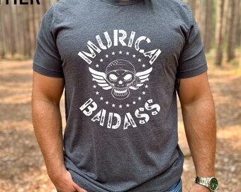 May include: Dark heather gray t-shirt with the text "Murica Badass" in white. The graphic features a skull wearing a bandana and wings, surrounded by stars. The shirt is a crew neck and has short sleeves.