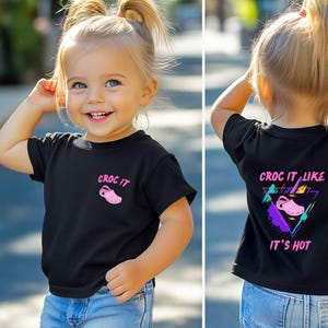 Könnte beinhalten: Ein schwarzes T-Shirt mit einer pinkfarbenen und blauen Grafik einer pinkfarbenen Clog und dem Text "CROC IT LIKE IT'S HOT". Das Shirt wird von einem Kind getragen.