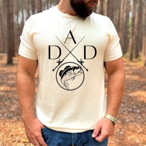 May include: A white t-shirt with a black graphic design of two crossed fishing rods with a fish jumping out of a circle. The graphic also includes the letters "A" and "D" on either side of the fishing rods.
