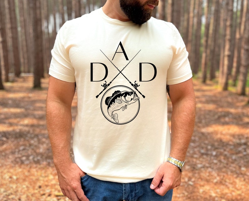 May include: A white t-shirt with a black graphic design of two crossed fishing rods with a fish jumping out of a circle. The graphic also includes the letters "A" and "D" on either side of the fishing rods.