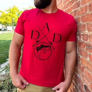 May include: Red t-shirt with a black graphic design of a fish and crossed fishing rods with the letters "DAD" around the design.