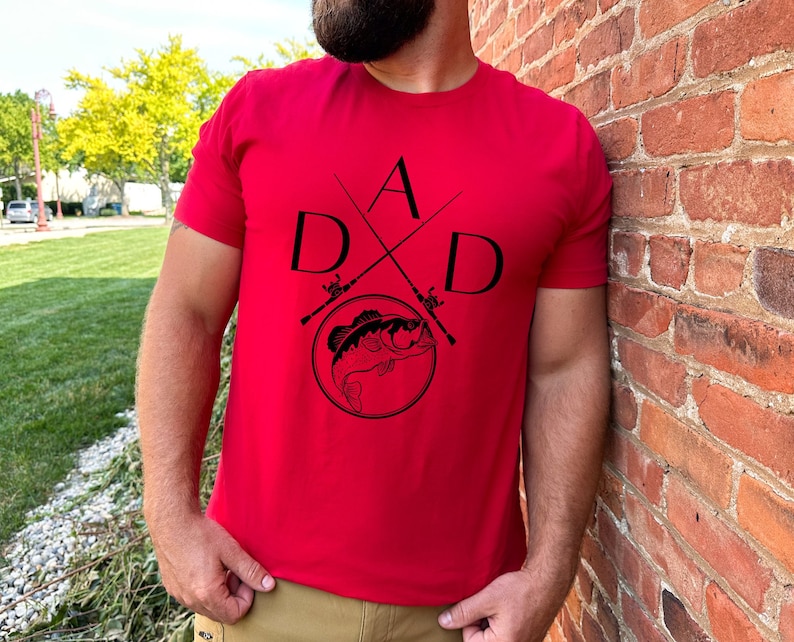 May include: Red t-shirt with a black graphic design of a fish and crossed fishing rods with the letters "DAD" around the design.
