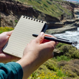 May include: A person holding a small, black, lined notebook with a black pen. The notebook has a spiral binding and the pen has the words "Caran d'Ache" printed on it. The person is standing on a cliff overlooking a rocky coastline with crashing waves.