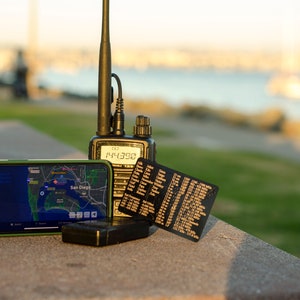 May include: A black handheld two-way radio with a telescoping antenna, a black smartphone displaying a map of San Diego, a black card with white text, and a black rectangular object on a gray concrete surface.