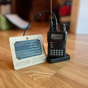 May include: A black handheld ham radio transceiver with a digital display showing the number 144390. The radio is sitting on a black charging base. Next to the radio is a brown card with the text "Ham Radio License Card" and the call sign KF6DT.