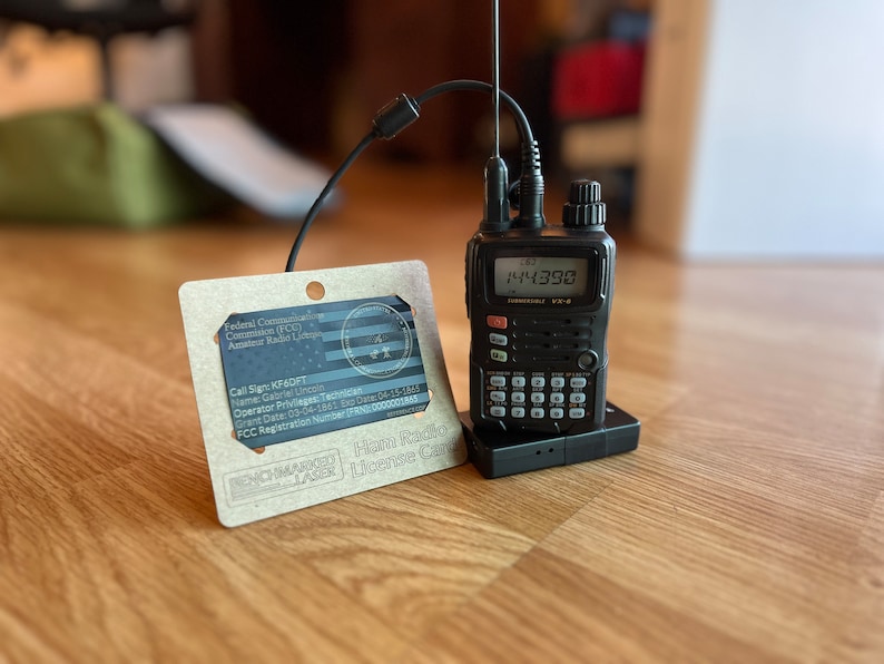 May include: A black handheld ham radio transceiver with a digital display showing the number 144390. The radio is sitting on a black charging base. Next to the radio is a brown card with the text "Ham Radio License Card" and the call sign KF6DT.