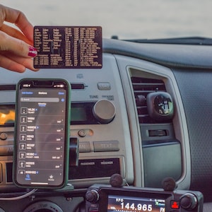 May include: A close-up of a car's dashboard with a mobile phone displaying a list of radio stations, a radio transceiver with a digital display showing "144.965" and "APRS", and a small card with a list of radio frequencies.