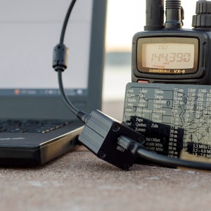 May include: A black handheld radio with a digital display showing "144.390" and the text "SUBMERSIBLE VX-6". The radio is connected to a black cable and a black box with the text "8837". A black and silver card with a map of the United States and frequency information is visible in the foreground.