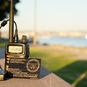May include: A black handheld two-way radio with a black antenna and a black cord. The radio has a digital display showing the number 144.390. A black and gold card with the text "Federal Communications Commission (FCC) Amateur Radio License" is next to the radio.