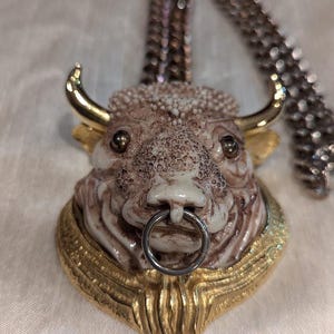 May include: A gold and white ceramic bull head pendant with a silver ring through the nose. The pendant is attached to a silver chain.