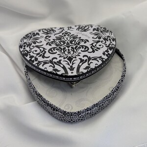 May include: Heart-shaped trinket box with a black and white damask pattern on the lid and sides. The box is open, revealing a white interior. The box is on a white fabric surface.