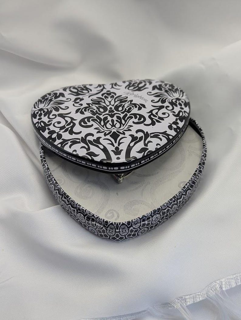 May include: Heart-shaped trinket box with a black and white damask pattern on the lid and sides. The box is open, revealing a white interior. The box is on a white fabric surface.