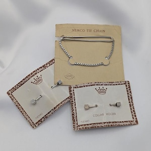 May include: Vintage NEMCO tie chain and collar holders on their original packaging. The tie chain is silver-toned with a chain design. The collar holders are small, silver-toned, and hexagonal. The packaging is tan with a crown design.