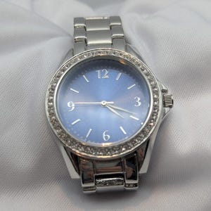 May include: A silver-toned wristwatch with a blue face and a bezel encrusted with small, clear crystals. The watch has a matching silver band and a crown on the right side. The watch face displays the numbers 12, 3, 6, and 9.