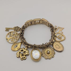 May include: Gold-toned charm bracelet featuring various charms, including a key, a tree, a leaf, a flower, a watch, and a heart-shaped charm. The watch has a white face and gold hands. The bracelet is a mix of chain and rectangular links.