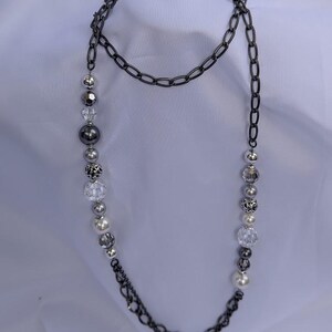 Puede incluir: Un collar largo con una cadena de metal de ca&ntilde;&oacute;n y una hebra de cuentas. Las cuentas incluyen esferas plateadas, transparentes y perladas. El collar se muestra sobre un fondo blanco, resaltando el contraste entre la cadena oscura y las cuentas m&aacute;s claras.