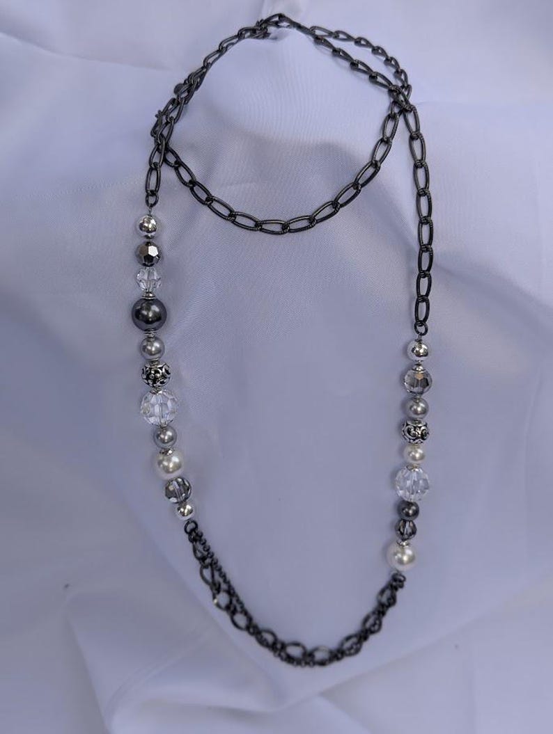 Puede incluir: Un collar largo con una cadena de metal de ca&ntilde;&oacute;n y una hebra de cuentas. Las cuentas incluyen esferas plateadas, transparentes y perladas. El collar se muestra sobre un fondo blanco, resaltando el contraste entre la cadena oscura y las cuentas m&aacute;s claras.