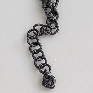 Puede incluir: Primer plano de una cadena de collar gris oscuro con un dije en forma de coraz&oacute;n. El dije tiene las iniciales "B" y "B". La cadena est&aacute; hecha de eslabones ovalados entrelazados y anillos redondos.