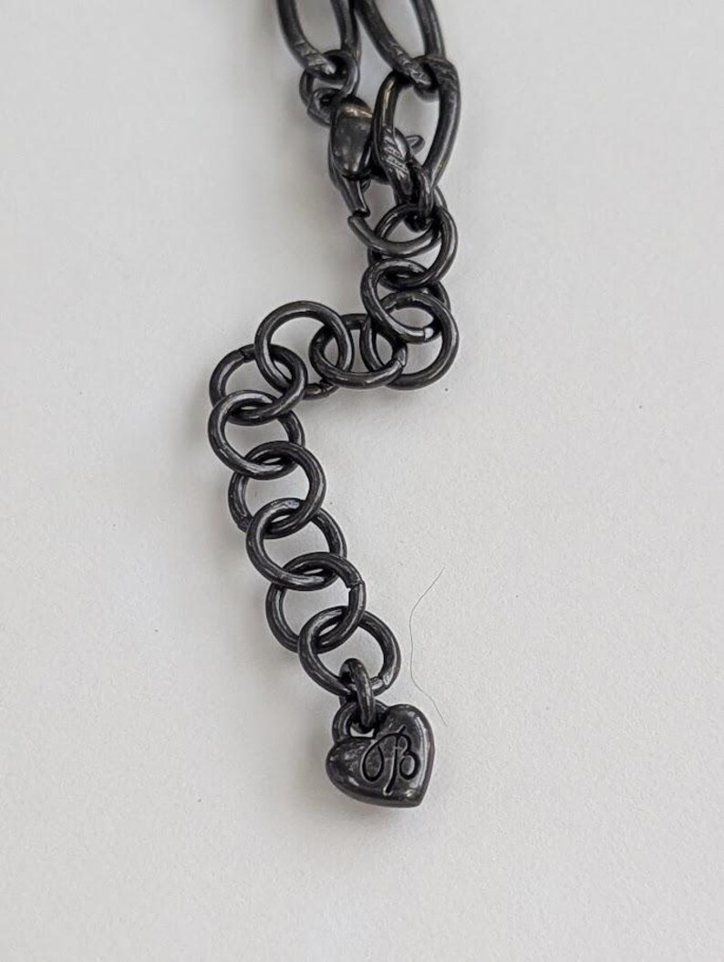 Puede incluir: Primer plano de una cadena de collar gris oscuro con un dije en forma de coraz&oacute;n. El dije tiene las iniciales "B" y "B". La cadena est&aacute; hecha de eslabones ovalados entrelazados y anillos redondos.