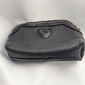 以下が含まれることがあります： ハート型の切り抜きがある黒い革の小銭入れ。表面は質感があり、上部にはダークブラウンのクロコダイル柄のトリムが付いています。小さなシルバー色のリングが側面に付いています。