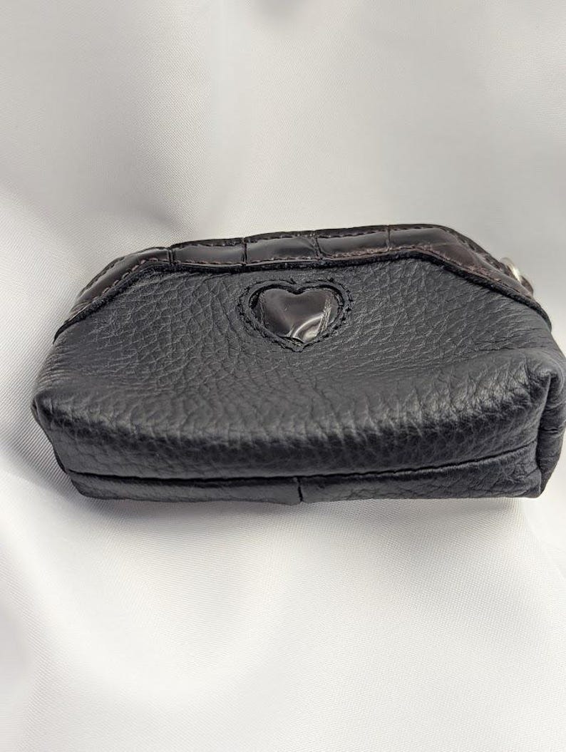 以下が含まれることがあります： ハート型の切り抜きがある黒い革の小銭入れ。表面は質感があり、上部にはダークブラウンのクロコダイル柄のトリムが付いています。小さなシルバー色のリングが側面に付いています。