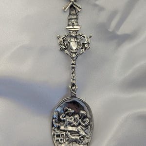 May include: A silver-colored spoon with a windmill design at the top. The spoon's bowl features a detailed relief scene of people gathered around a table. The handle has ornate scrollwork and a heart-shaped element.