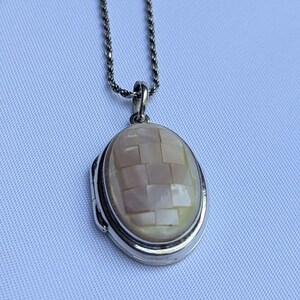 May include: Oval silver pendant necklace with a mosaic-style mother-of-pearl inlay. The pendant is suspended from a delicate silver chain. The locket design adds a classic touch to this jewelry piece.