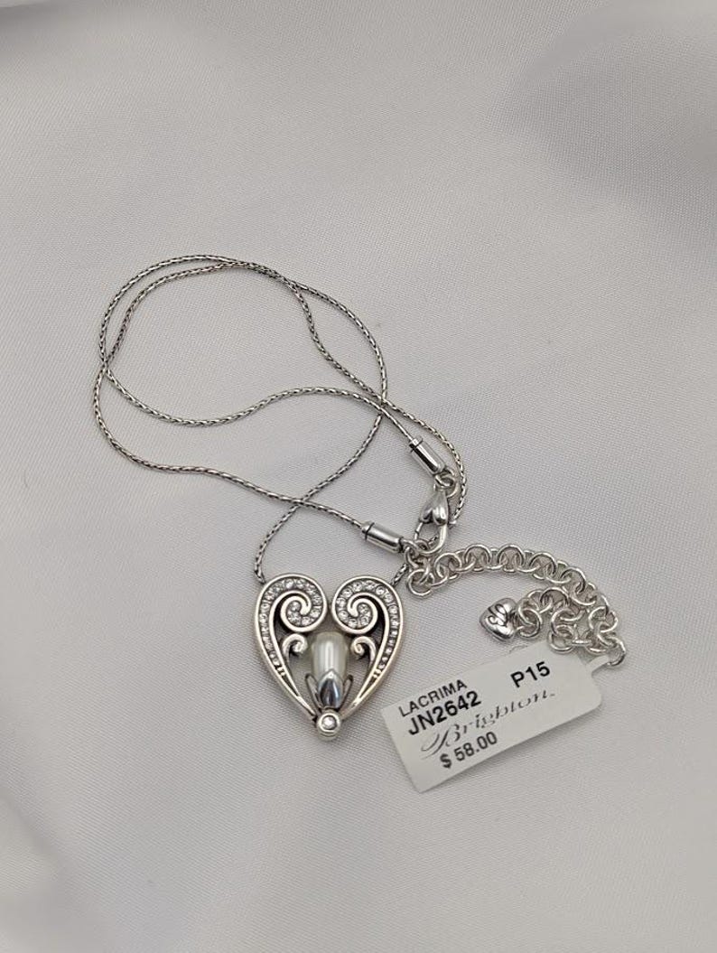 May include: Silver heart pendant necklace with a pearl-like centre and clear crystal accents. The heart design features swirling details and is suspended from a delicate chain. A tag with the text "LACRIMA JN2642 Brighton" is attached.