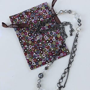 Puede incluir: Un collar plateado con una mezcla de cuentas y eslabones de cadena, junto a una bolsa con estampado floral. El collar presenta cuentas transparentes, grises y perladas. La bolsa tiene una cinta marr&oacute;n con la palabra "Brighton" impresa.